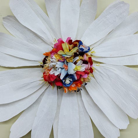A giant paper daisy, created by artist Ling Warlow and local school children. White outer ray florets encircle a brightly coloured composite centre with the disc florets in an array of colours and flower shapes