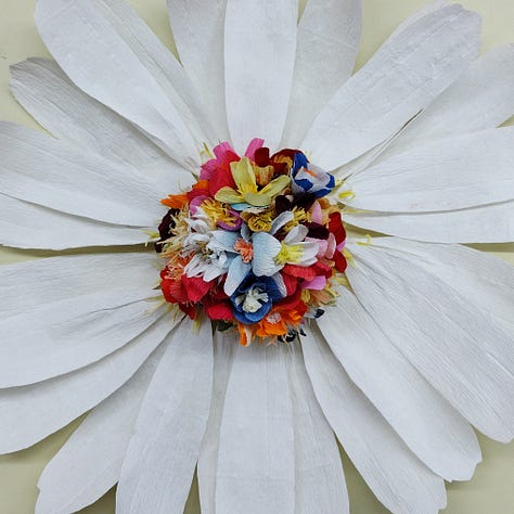 A giant paper daisy, created by artist Ling Warlow and local school children. White outer ray florets encircle a brightly coloured composite centre with the disc florets in an array of colours and flower shapes