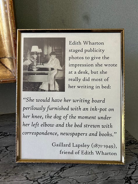 1: Informational display titled "The Nature of Houses" with text and decorative motifs. Transcribed Text: The Nature of Houses In her fiction Wharton often drew parallels between houses and human character, most notably in her 1892 short story, "The Fulness of Life": "I have sometimes thought that a woman’s nature is like a great house full of rooms: there is the hall, through which everyone passes; the drawing room, where one receives formal visits; the sitting room, where the members of the family come and go . . . but beyond that, are other rooms, the handles of whose doors are never turned; no one knows the way to them, no one knows whither they lead; and in the innermost room, the holy of holies, the soul sits alone and waits for a footstep that never comes." She later critiqued this semi-autobiographical depiction of a soul in pain as "one long shriek [written]  at the top of my voice." 2: Elegant library with dark wood and floor-to-ceiling bookshelves, ornate decorative features, antique furniture, and large windows. 3: Framed image of Edith Wharton sitting at a desk with a text quote beside her portrait. Transcribed Text: Edith Wharton staged publicity photos to give the impression she wrote at a desk, but she really did most of her writing in bed: “She would have her writing board perilously furnished with an ink-pot on her knee, the dog of the moment under her left elbow and the bed strewn with correspondence, newspapers and books.” Gaillard Lapsley (1871-1949), friend of Edith Wharton. 4: Picture of an elegant dining room with a round table covered by a white tablecloth, set with crystal glasses and candlesticks. The floor is carpeted in a beige, woven pattern. Under the table, a small, fluffy dog with long hair is lying on a leopard-print cushion. 5: Weathered gravestone in a forest with the inscription "Our Sweetheart Modele, Died Jan 5 1914." 6: Poster with a photo of an interior hallway and text about the purpose of doors. Transcribed Text: Above all, privacy For Wharton, doors created privacy as well as beauty. Inspired by European houses, the main floor of The Mount is in a style called enfilade. All rooms open up to the next room and doors are lined up to allow vistas from one room to another. “…the main purpose of a door is to admit, its secondary purpose is to exclude.”  Edith Wharton, The Decoration of Houses, 1897 7: Photograph depicting white and pink flowers, which circle around a small man-made pond. In the background, a manicured garden is visible. 8: An open spiral notebook with handwritten notes on the right page. Transcribed Text: YES AGREE! More fake dogs please! yes ma'am! yess! please!! Edith has such a slay style! Love her work in both design, art & literature! ~ Kloey More fake dogs plz! ❤️[heart] 9: Text on a beige wall about Edith Wharton's writing style. Near the left side, the text reads, "The innermost room..." in a larger, bold serif font. Below this, there is a paragraph in a smaller, sans-serif font describing the writing style of Edith Wharton.  Transcribed Text: "The innermost room..." Edith Wharton did not write happy endings. Her stories feature the lost chance; her characters yearn for someone to join them in that innermost room of the soul. Whether Wharton achieved that in her own life, and with whom, is a story as mysterious and captivating as any of her fiction.