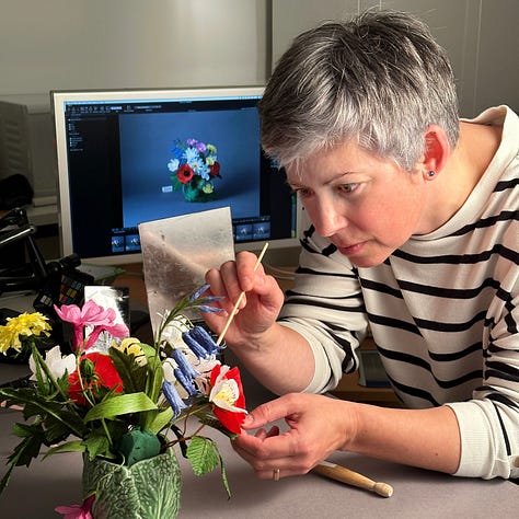 A beautiful arrangement of wildflowers in bright shades of pink, yellow, red, blue and white, presented in a green cabbage leaf mug, against a dark grey background. Two pictures of the author trying not to laugh, or breathe too hard, on the arrangements as she makes the final tweaks!