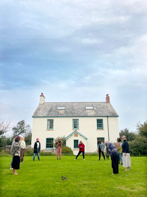 L-R: three white women and one Black woman sit on sofas and the floor writing in journals, with tissues, herbs and a jug of herbal tea in the foreground. A group of nine women stand in a circle on the lawn of a farmhouse, in the background. 10 women eating lunch around a farmhouse style table, with floral arrangements in the centre. Three women crouch on the wooden floor of a barn, their backs to us, lighting candles. In the foreground is a floral mandala arrangement. Four women clamber down a steep coastal path to the beach