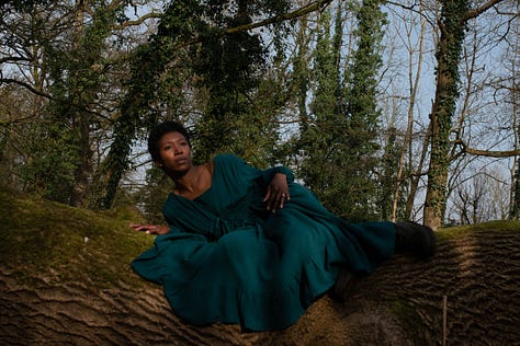 A Black woman with short afro hair wears a long, teal dress. She is reclining on moss covered fallen tree in shadow and sunlight against a backdrop of blue sky and ivy wrapped tree trunks.