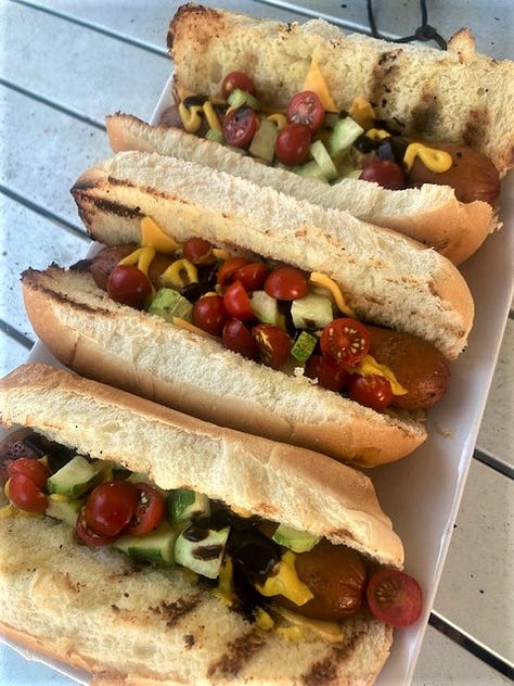 Grilled hotdogs being prepared on an outdoor grill with a number of ingredients and the buns.