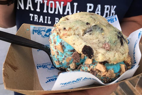 Three images of large delightful cookies with blue ice cream in the middle and toffee chunks or sprinkles around the outside edges of the ice cream, all being held in wrappers that say The Baked Bear.