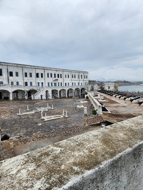 A model of Cape Coast Castle with white stone walls, and red roofing, An outdoor image on the castle grounds showing white stone and concrete pavement. An inscription in the castle wall entitled "In Everlasting Memory."