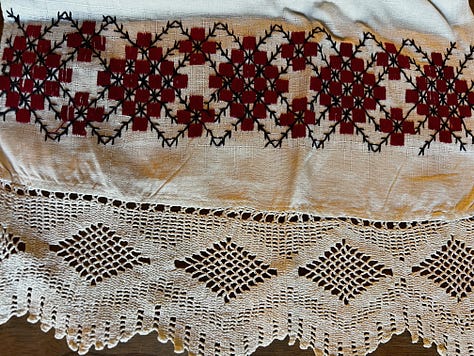 Three photos of embroidery work. Left, a white linen tablecloth edged with lace and embroidered with a geometric pattern in red; in the middle a kind of cotton net sewn with decorative patterns in red, yellow, and green yarn with "1906" in the middle; on the right, a white linen table runner embroidered with a red bird and black decorative edging.