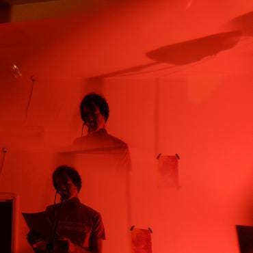 Three images of Stu reading in a room lit by red lights. Location: The Auxiliary Project, Middlesborough. July 2023