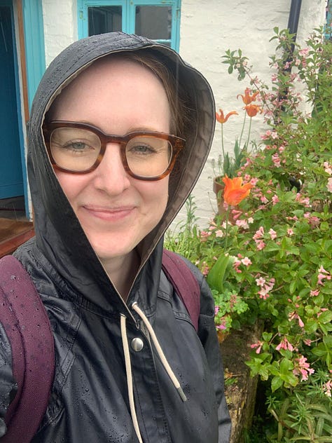 L-R: pink flowers, Emily (a white woman wearing glasses) wears a raincoat, hood up, taking a selfie in front of wildflowers, a door with "self-service pottery shop" written on it