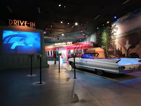 Panorama of display inc,luding huge convertible car and drive-in movie screen, huge convertible and poster advertising freeway travel,