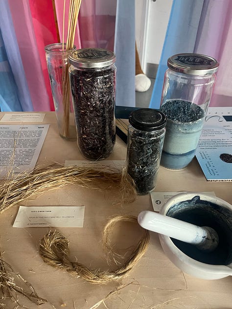 1-3. Exhibition table for 'garden indigos' from Indigo Garden and Fantasy Fibre Mill, showing dyed indigo fabric, processed linen fibre, woad seeds, Japanese indigo pigment, dried Japanese indigo leaves, retted and hackled flax straw and information boards.