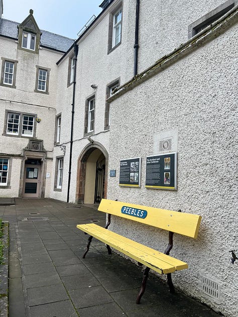 a bright yellow wooden bench with Peebles on a metal sign sits in the Quadrange off Peebles high street near the war memorial.