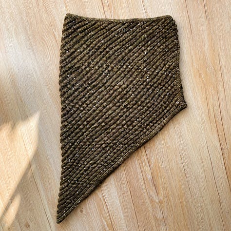 A brownish-green knitted cowl laying on a light brown floor in different positions.