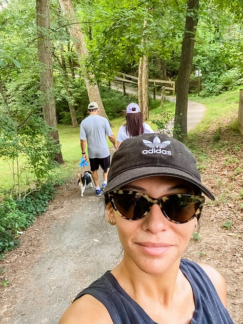 Photo of a paved walking trail surrounded by lush vegetation and a deer on one side, a round embroidery hoop with an image of a pineapple, a woman smiling and presenting the piles of fresh pasta she made from scratch, a selfie photo of woman and her dad painting a piece of furniture, a photo of a woman and her mom sitting on the floor painting a piece of furniture, a selfie photo of a woman and her parents sitting down at the dinner table to eat barbecue food, a photo of a woman and her dog looking out the window, a photo of a LOVE sign in a grassy, woody green forest, a selfie photo of a woman with her parents and dog behind her walking in the opposite direction.