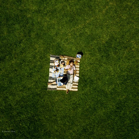 A grid of six images showing scenes from the "Powers of Ten" film. Top row, left to right: A close-up of two people lying on a picnic blanket with various items around them; a more zoomed-out view showing them as a small spot on a large expanse of grass; and a view of Earth from space. Bottom row, left to right: a sequence of images showing views at different scales, starting from Earth and zooming in closer until individual objects and patterns appear; a close-up of a man lying on the picnic blanket; and the title screen reading "Powers of Ten."