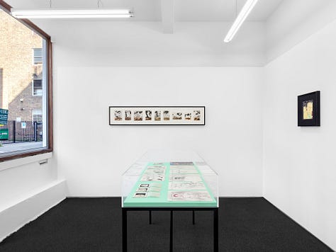 1) The table full of works is in the center of the image. It has a black frame and a clear top. There are also several frames of Sadao's work hanging on all the surrounding walls. 2) A straight down view of a table is seen showing many different drawings from Sadao. There are various sketches, and a cover of a gay magazine. 3) A clear showcase table is in the middle of a room, with a window to the left. There is a long horizontal frame on the wall full of Sadao's work on the back wall, and another black framed image on the right wall. The table in the center is full of his drawings.