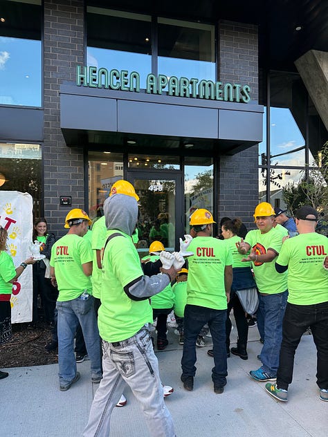 construction workers wearing yellow hats and neon green shirts stand in front of the entrance to an apartment building, a banner reading "nothing gets built without these hands" stands in front of a few dozen white plaster mold hands, and a poster board of an article stands next to a cardboard cut out of a construction worker