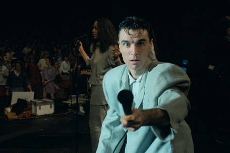 selects from Talking Heads' concert film Stop Making Sense. images of Byrne holding a microphone to camera, raising his arms, dancing in a big suit an with a lamp and full shots of the band.