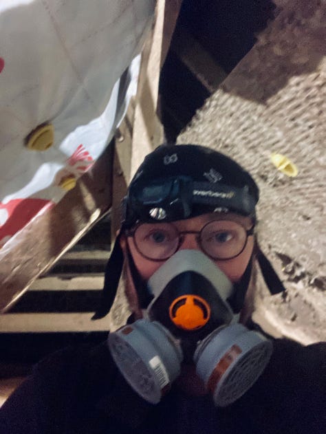 Steve in a breathing mask, glasses, and a black bandana over his hair looks out at us from the top of the scaffold in this selfie. His head is at the apex of the partly insulated roof. 