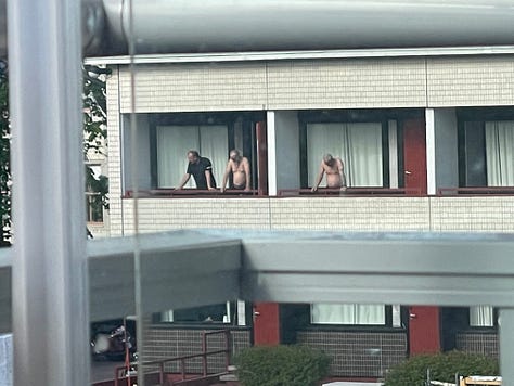 A two story building in beige with a dark grey roof form 2 angles. A group of shirtless older men standing on their balcony looking out into my balcony