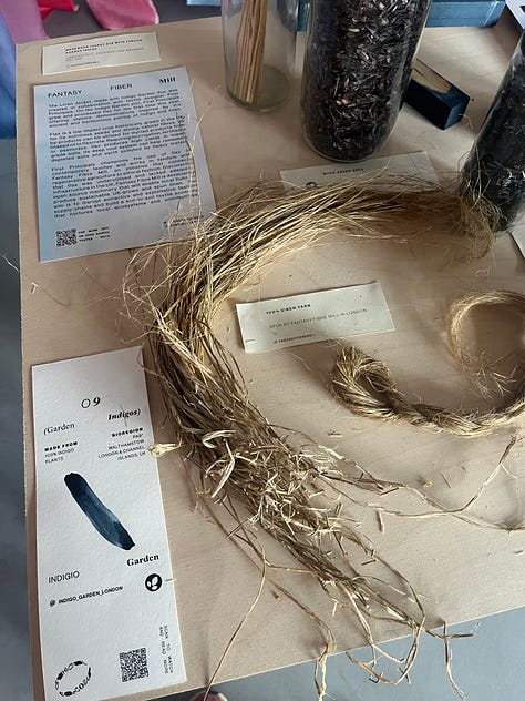 1-3. Exhibition table for 'garden indigos' from Indigo Garden and Fantasy Fibre Mill, showing dyed indigo fabric, processed linen fibre, woad seeds, Japanese indigo pigment, dried Japanese indigo leaves, retted and hackled flax straw and information boards.