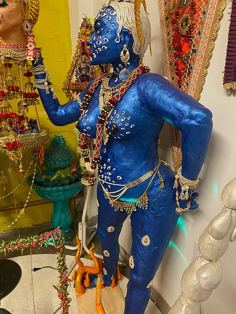 Blue goddess statue, working class woman and her daughter both in pink hijabis, gold-colored goddess icon with orange detail.