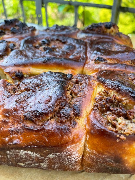 Golden-baked spiced sweet rolls nestled in a parchment-lined square tin, their glossy tops richly browned and dotted with caramelized raisins and nutty filling. One roll is pulled slightly away, revealing a soft, fluffy interior marbled with rum-soaked raisins and a fragrant, spiced nut mixture. Close-up shots highlight the tender, airy crumb and the dark, buttery swirl of filling, contrasting beautifully with the deeply bronzed crust. The setting hints at a cozy, sunlit kitchen or balcony, adding warmth to this homemade treat.