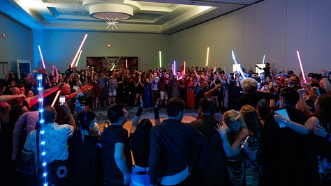 1: Cass on stage with the Halcy-Con event team, all arm-in-arm | 2: A massive circle of people hugging and swaying to a song; Cass is visible in the foreground, definitely trying to hide the fact that she's weeping by tucking her head in a friend's shoulder | 3: Cass beneath an arch of lightsabers, hand held to her mouth because there's really no hiding that she's crying this time.