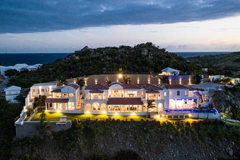 An aerial view of Villa Amalia perched on the cliffs of Oyster Pond, St. Martin, showcasing its sleek modern design with an expansive infinity pool overlooking the turquoise Caribbean Sea. The villa’s open-concept layout, floor-to-ceiling glass walls, and spacious outdoor terrace offer a perfect blend of indoor and outdoor luxury living, with lush greenery surrounding the property and the ocean visible in the distance.