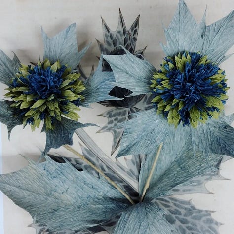 Image one depicts sea holly made from paper dyed with a ranof natural colours, 8mage 2 shows the natural plant growing in its natural beach environment, spiky, holly shaped leaves of a glowing, pale blue-green hue. Picture 3 shows students in the art studio holding their paper sea holly and smiling