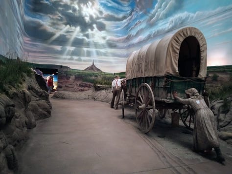 Three views of same tableau: Overview, showing a life-sized covered wagon being pulled by oxen and pushed from behind by woman in dress, as it heads toward the spire of Chumney Rock, a major landmark, and the sun sets. Close up of woman pushing, showing Oregon or Bust painted in large letters on side of covered wagon; view from front, showing man in hat cracking whip over four oxen pulling wagon