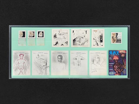 1) The table full of works is in the center of the image. It has a black frame and a clear top. There are also several frames of Sadao's work hanging on all the surrounding walls. 2) A straight down view of a table is seen showing many different drawings from Sadao. There are various sketches, and a cover of a gay magazine. 3) A clear showcase table is in the middle of a room, with a window to the left. There is a long horizontal frame on the wall full of Sadao's work on the back wall, and another black framed image on the right wall. The table in the center is full of his drawings.