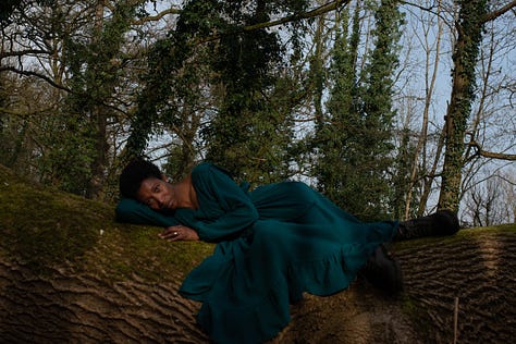 A Black woman with short afro hair wears a long, teal dress. She is reclining on moss covered fallen tree in shadow and sunlight against a backdrop of blue sky and ivy wrapped tree trunks.