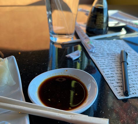 glass of water next to a bottle of soy, table top with saucer and napkin written on, plate of sushi in foreground