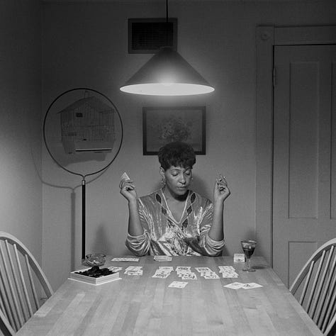 1-9) All images are black and white, and involve a table, as well as a few chairs, and an overhead light. 1) A black woman is seen playing cards by herself. 2) A black woman is seen holding her head in her hands, while reading a book. There is a child sitting to her right, writing. 3) A black person is standing at the end of the table, looking into the camera. 4) A black woman is looking at herself in a tabletop mirror, while a child is playing to the right of her at the table. 5) Three people are sitting at the table, and appear to be playing a game or laughing. 6) A black woman is seen looking at herself in a tabletop mirror. 7) A black woman is sitting on the left side of the table, with her hand under her chin. There is a black man at the end of the table, reading the newspaper, with his hand resting on his neck. 8) Two black people are seen kissing/embracing. One is sitting at the end of the table, and the other person is behind them. 9) A black woman is seen with her head laying back on the table, with her hands in her hair.