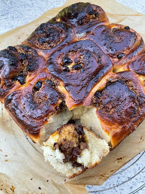 Golden-baked spiced sweet rolls nestled in a parchment-lined square tin, their glossy tops richly browned and dotted with caramelized raisins and nutty filling. One roll is pulled slightly away, revealing a soft, fluffy interior marbled with rum-soaked raisins and a fragrant, spiced nut mixture. Close-up shots highlight the tender, airy crumb and the dark, buttery swirl of filling, contrasting beautifully with the deeply bronzed crust. The setting hints at a cozy, sunlit kitchen or balcony, adding warmth to this homemade treat.
