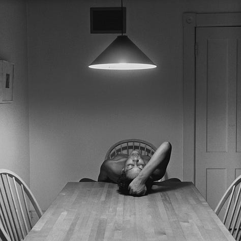 1-9) All images are black and white, and involve a table, as well as a few chairs, and an overhead light. 1) A black woman is seen playing cards by herself. 2) A black woman is seen holding her head in her hands, while reading a book. There is a child sitting to her right, writing. 3) A black person is standing at the end of the table, looking into the camera. 4) A black woman is looking at herself in a tabletop mirror, while a child is playing to the right of her at the table. 5) Three people are sitting at the table, and appear to be playing a game or laughing. 6) A black woman is seen looking at herself in a tabletop mirror. 7) A black woman is sitting on the left side of the table, with her hand under her chin. There is a black man at the end of the table, reading the newspaper, with his hand resting on his neck. 8) Two black people are seen kissing/embracing. One is sitting at the end of the table, and the other person is behind them. 9) A black woman is seen with her head laying back on the table, with her hands in her hair.