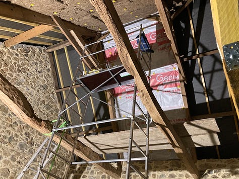 Steve in a breathing mask, glasses, and a black bandana over his hair looks out at us from the top of the scaffold in this selfie. His head is at the apex of the partly insulated roof. 