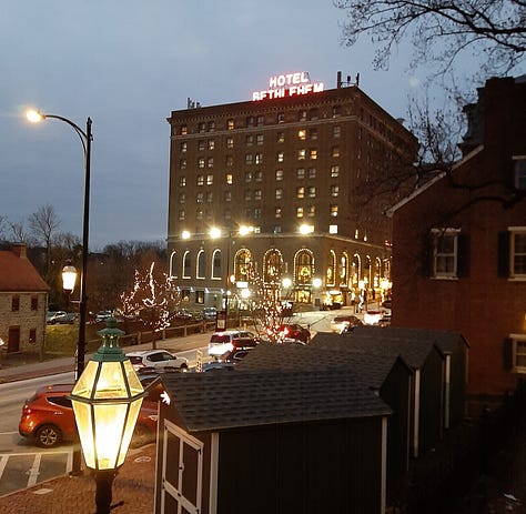 A collage of photos showing the Historic Hotel Bethlehem, including two photos of the 9-story brick exterior, and interior pictures of the elevators, white marble staircase with polished handrail, open doors to the ballroom with a glistening chandelier, and two photos of the lobby, with luxurious black leather furniture in front of a huge arched window with a Christmas wreath in the middle. Also a look inside a room of a bed with a robe lying on the end.