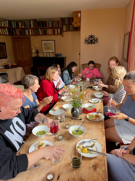 L-R: three white women and one Black woman sit on sofas and the floor writing in journals, with tissues, herbs and a jug of herbal tea in the foreground. A group of nine women stand in a circle on the lawn of a farmhouse, in the background. 10 women eating lunch around a farmhouse style table, with floral arrangements in the centre. Three women crouch on the wooden floor of a barn, their backs to us, lighting candles. In the foreground is a floral mandala arrangement. Four women clamber down a steep coastal path to the beach