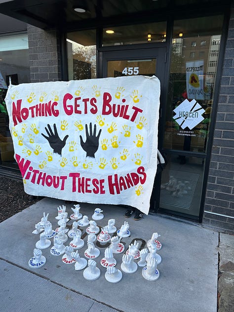 construction workers wearing yellow hats and neon green shirts stand in front of the entrance to an apartment building, a banner reading "nothing gets built without these hands" stands in front of a few dozen white plaster mold hands, and a poster board of an article stands next to a cardboard cut out of a construction worker