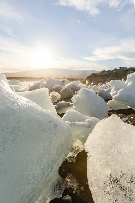 Photo of woman smiling over her shoulder sitting atop a boulder and wearing winter clothes, photo of a white and red church atop a green and yellow hill, photo of a woman wearing winter clothes smiling sitting on a boulder along a glacial lagoon river with large chunks of ice, photo of a small tow along a green coast with a jagged green and brown mountain rising up from the coastal edge in the background, photo of a woman standing on a boulder with a waterfall in the background, photo of a jagged green and gray rocky canyon with a river winding through, photo of large chunks of glacial ice on a beach, photo of two men and two women staggered on a rainbow-colored road that leads to a church, photo of the interior of a diner with a red cooler and a woman serving coffee