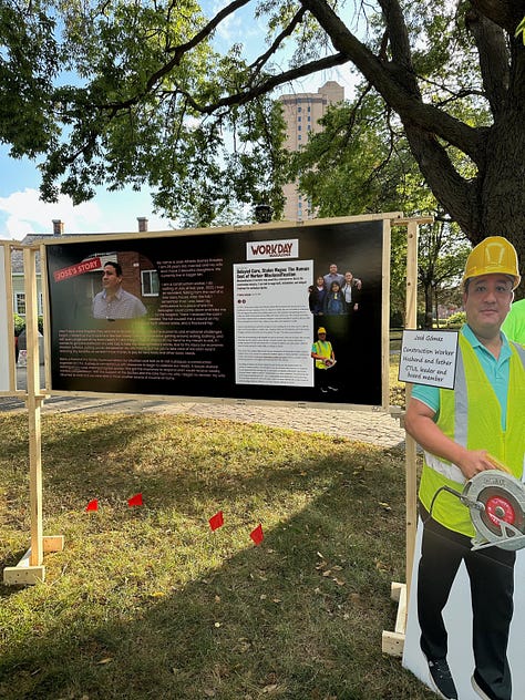 construction workers wearing yellow hats and neon green shirts stand in front of the entrance to an apartment building, a banner reading "nothing gets built without these hands" stands in front of a few dozen white plaster mold hands, and a poster board of an article stands next to a cardboard cut out of a construction worker