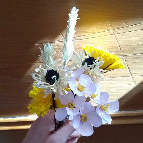 Image one is a hand holding a collection of rather clumsy flowers made from tissue paper, one recognisably a ragged robin, the others yellow and white. Image tow is a hand holding a slightly more refined bunch of crepe paper flowers, dandelion, plantain, cuckoo flower and grasses. The third image is an elegant stem of pure white meadow cranesbill, made of paper