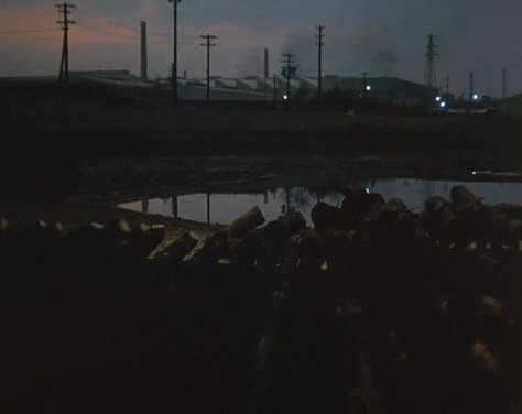 Nine screengrabs from AKA Serial Killer. They show in order: A Japanese street by day, with a car. A Japanese street at dusk, with lights reflecting on the wet gound. A military march, with the Imperial Japanese Army/Rising Sun flag, the Japanese state flag, and a third military flag I can't identify. A railway station with two unmoving wagons. A woman lying in a man's lap in a park by daylight. A car parkwith many grey cars. A sign in Japanese and English, the English reading "United States Forces Japan Area Unauthorized Entry Punishable By Japanese Law". A pile of felled trees at dusk, with a factory building across the river smoking in the background. A street by day with many cars driving upwards as if into the sky and one person on a bike coming down the hill.