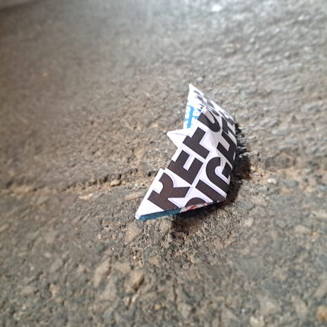 an origame paper boat left on the path. paper had 'refugee rights' written in block print across it.