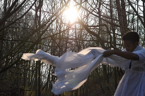 Me, a Black woman wearing a floaty white top is dancing with a white voile which billows around her. The weak Spring sunlight flares through the trees in the woodland behind her. 