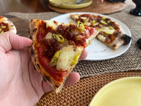 Bowls of peppers, cheese, sauce and txistorra, the pizza out of the oven, a hand holding a slice of the pizza
