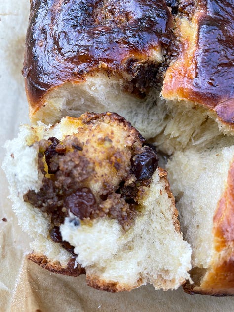 Golden-baked spiced sweet rolls nestled in a parchment-lined square tin, their glossy tops richly browned and dotted with caramelized raisins and nutty filling. One roll is pulled slightly away, revealing a soft, fluffy interior marbled with rum-soaked raisins and a fragrant, spiced nut mixture. Close-up shots highlight the tender, airy crumb and the dark, buttery swirl of filling, contrasting beautifully with the deeply bronzed crust. The setting hints at a cozy, sunlit kitchen or balcony, adding warmth to this homemade treat.