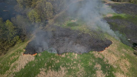 Fire burns in an open grass field, photo of fire and smoke from above, a person in orange jumpsuit walks up a blackened hill