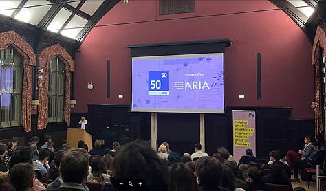 1. A photo of a sign saying 'Cambridge Neuroworks, Powered by ARIA, Cambridge and the Advanced Research + Invention Agency (ARIA). Accelerating the translation of ideas to find revolutionary neurotechnologies. There are people mingling in the background of the image. 3. A photo of a person presenting to a room full of people, there is a screen in the background saying 'Powered by ARIA' and the Fifty Years logo on it. 4. A photo of people mingling at an event. In the background there is a large vinyl decal saying 'Hello brain!' 5. A room full of people listening to someone speak (off camera) at a Pillar VC event. 6. A person presenting to a room of people. 